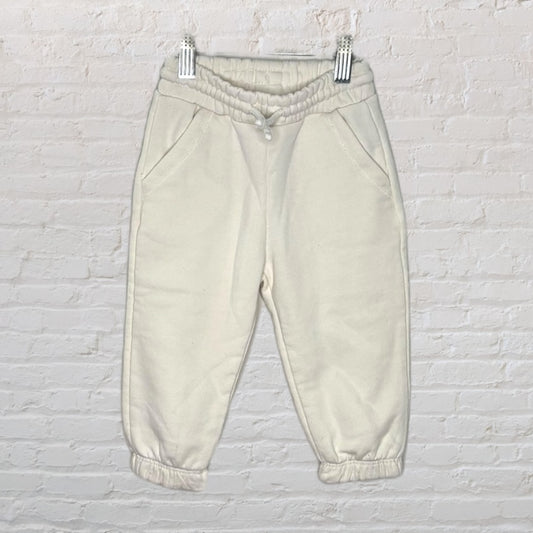 Light beige casual jogger pants with elastic waistband and cuffs, hanging on clips against a brick wall background.