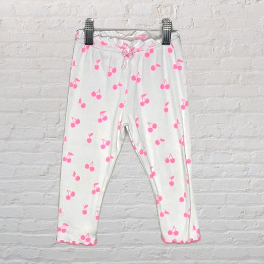 White pants with pink cherry print, featuring an elastic waistband and ruffled hem, hanging on a white wall.