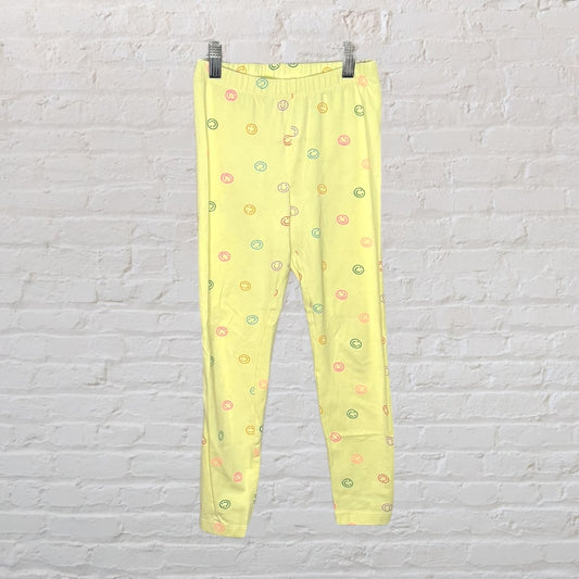 Yellow leggings with colorful smiley faces printed, displayed against a white brick wall.