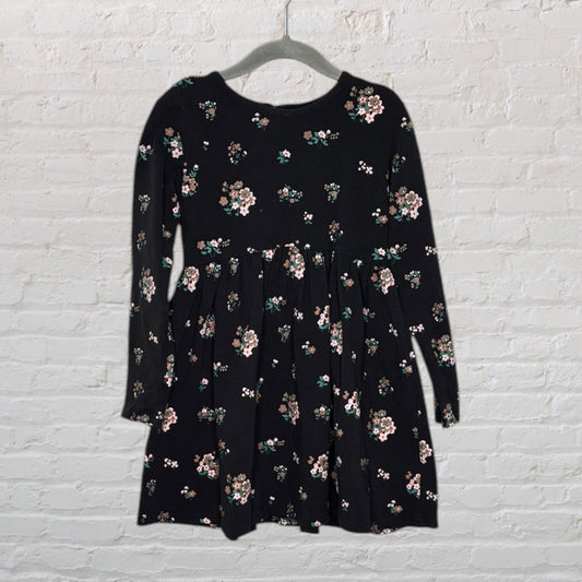 Black long-sleeve dress featuring a floral pattern, displayed on a hanger against a brick wall.