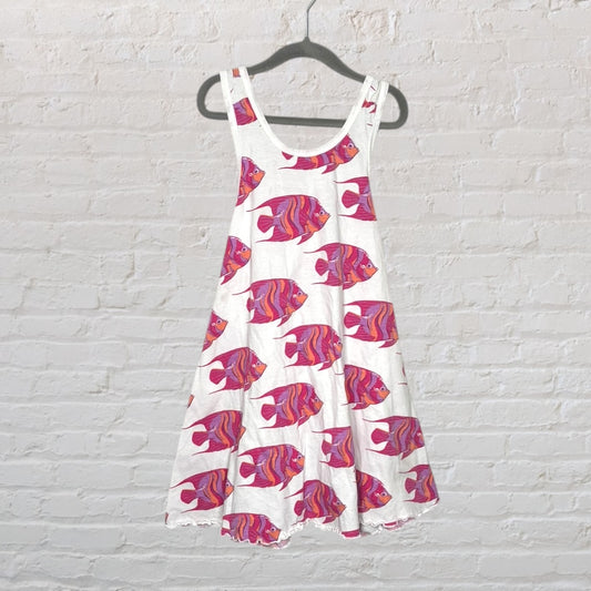 White sleeveless dress with pink fish print, hanging on a hanger against a brick wall.