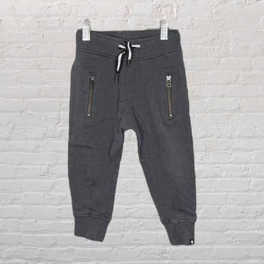 Children's charcoal gray jogger pants with zipper pockets and drawstring waist, displayed against brick wall.