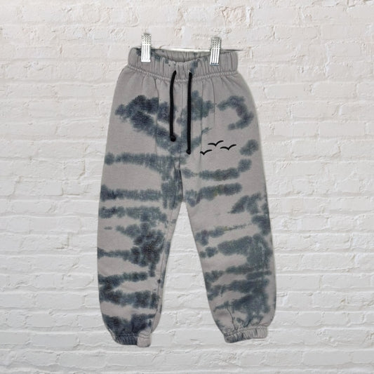 Gray tie-dye sweatpants with black drawstring and bird motif, displayed against a white brick wall.