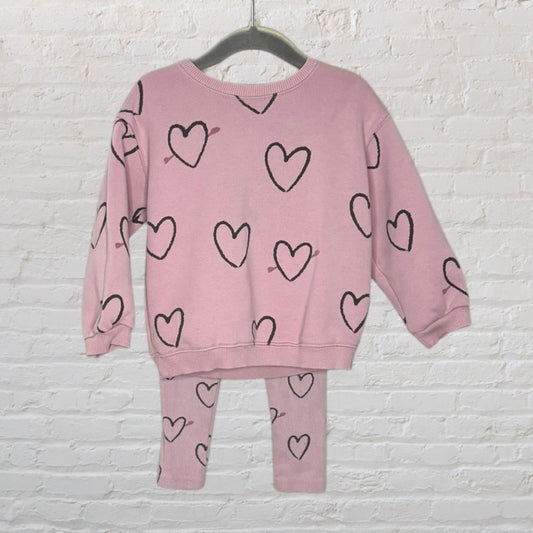 Pink sweater and leggings set with black heart pattern, displayed on a hanger against a white brick wall.