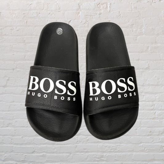 Black Hugo Boss slide sandals with white logo text on a white brick background.