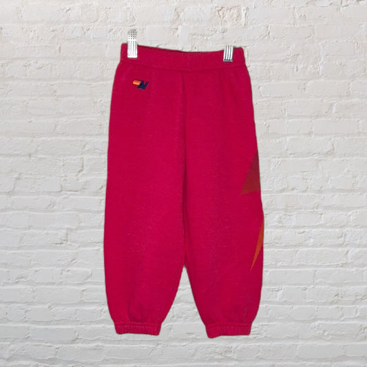 Bright pink sweatpants with a lightning bolt graphic on the side and elastic cuffs, hung against a white brick background.