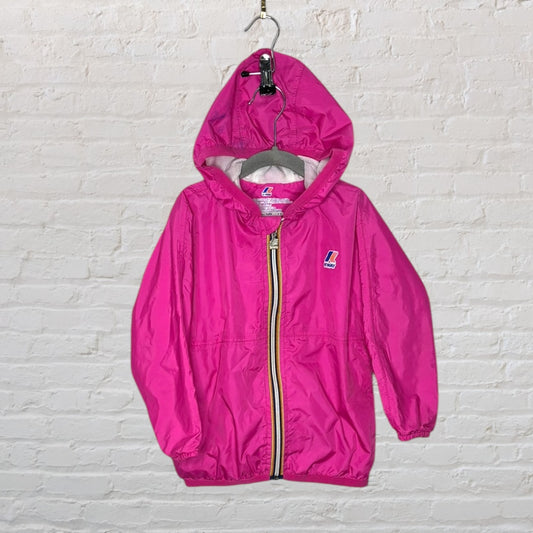 Pink hooded windbreaker jacket by K-Way, displayed on a white brick wall background.