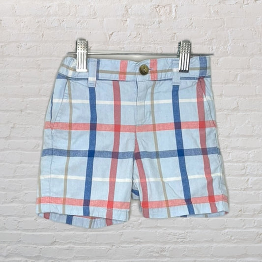 Unisex plaid shorts featuring light blue, red, and beige colors, displayed on clip hangers against a white brick wall.