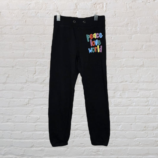 Black sweatpants with 'peace love world' multicolour text on the left side, hanging on white brick background.